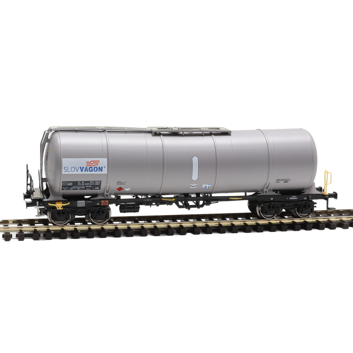 Grey four-axle Slovvagon Zacns tank wagon model in TT scale on a realistic modern industrial railway layout, oil depot with storage tanks in the background, soft daylight, highly detailed miniature scene
